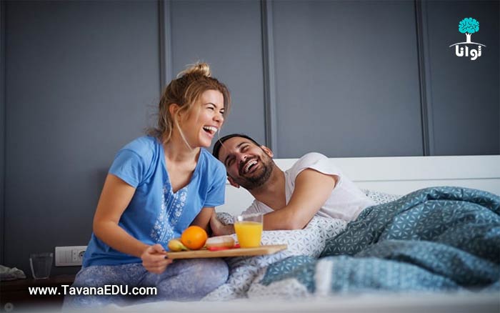 زن و شوهری روی تخت خواب در حال صبحانه خوردن و خندیدن